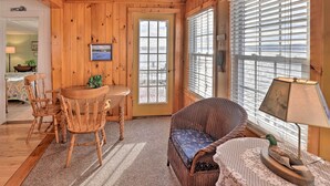 Une pièce chaleureuse aux murs en bois, avec une table à manger et des chaises, un fauteuil confortable, une lampe et une fenêtre équipée de stores.