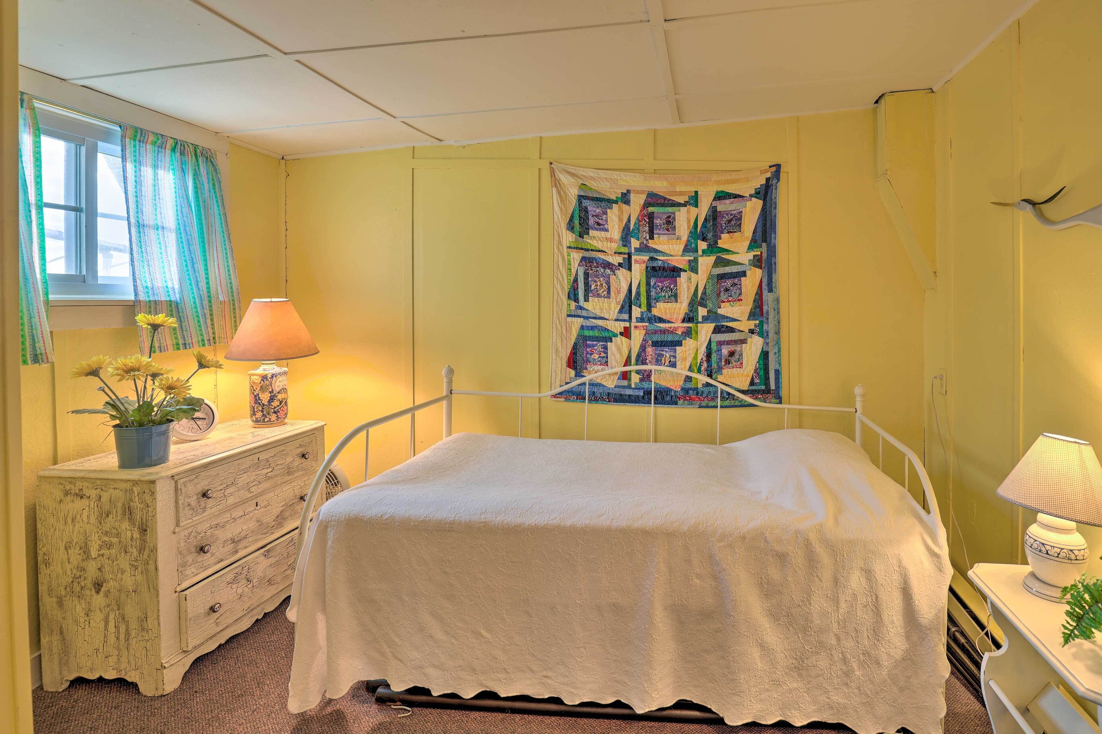 A bedroom with a bed, a dresser, a lamp, and a quilt on the wall.