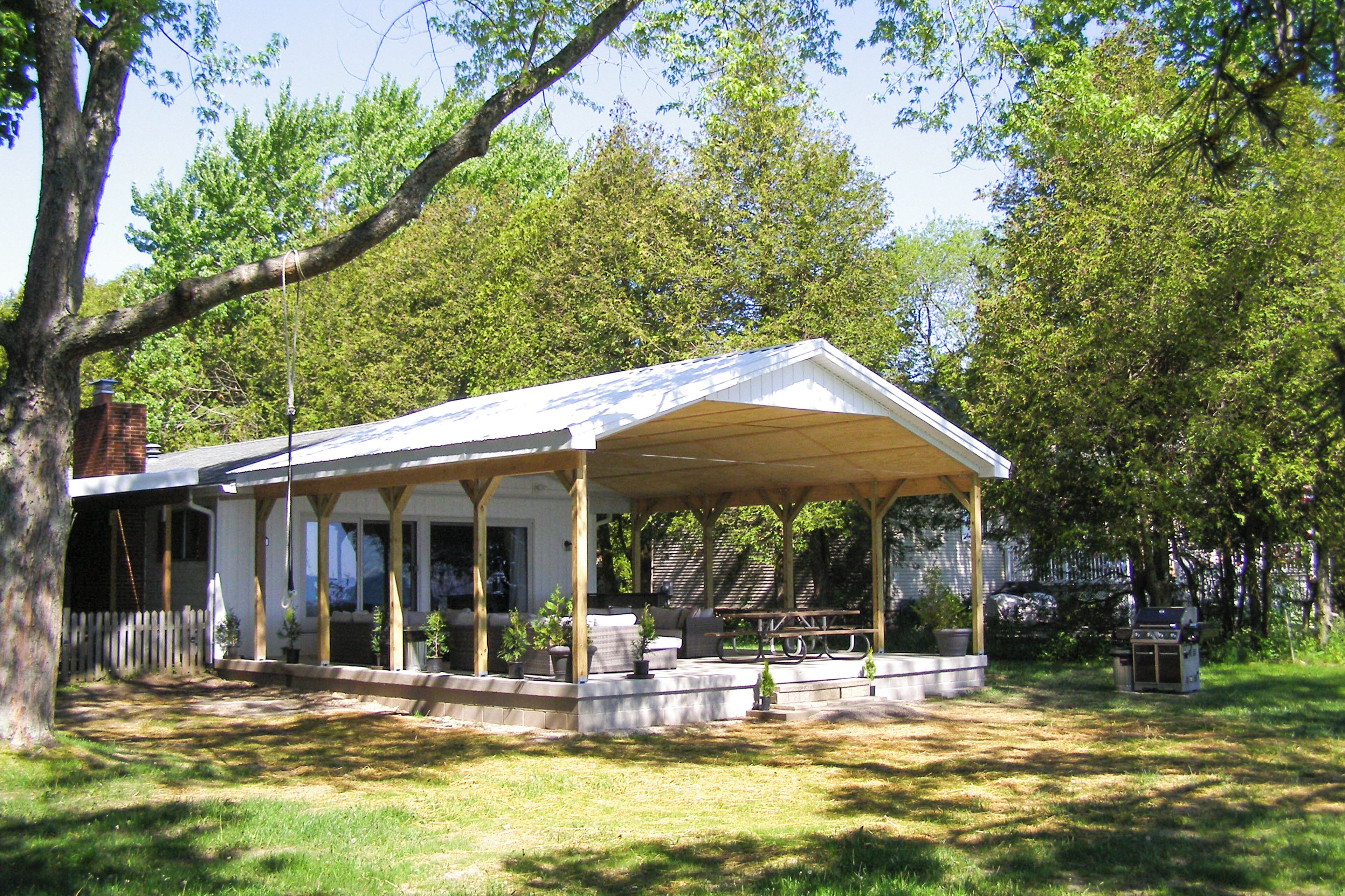 A covered outdoor seating area with a table and chairs, surrounded by potted plants and a barbecue grill.