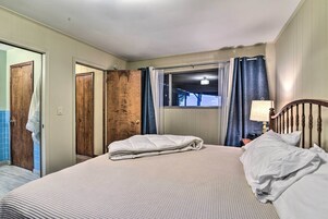 Una cama bien hecha con edredón blanco y almohadas, cabecera de madera, ventana con cortinas y una lámpara en la mesita de noche.