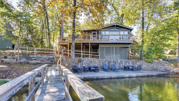 Eine Holzterrasse führt zu einem Haus mit einem Balkon, der auf einen Teich mit Baumumrandung blickt.