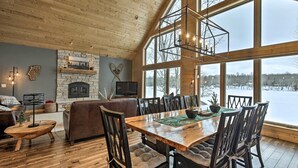 A spacious dining area with a wooden table and chairs, a stone fireplace, and large windows offering a view of the outdoors. - Lakefront Cottage in Iron River: 3 Mi to Skiing! (Iron River)