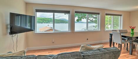 A living room with a gray sofa, a flat-screen TV mounted on the wall, and a dining area with a table and chairs. Large windows provide a view of a lake and distant buildings.