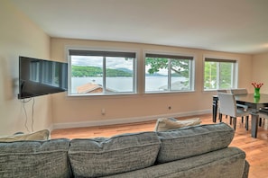 Sala de estar con un sofá gris, un televisor de pantalla plana montado en la pared y un comedor con mesa y sillas. Amplias ventanas ofrecen vistas a un lago y edificios lejanos.