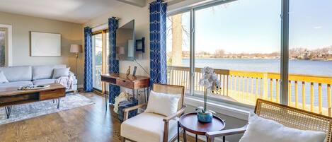 A spacious living room with a large window overlooking a body of water, a sofa, a dining table, and a chair.