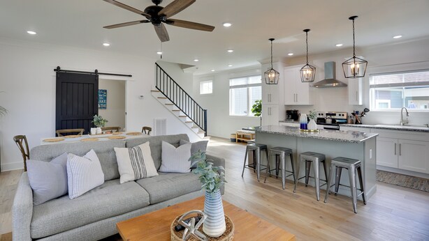 A spacious living area with a gray sofa, wooden coffee table, and a ceiling fan. A kitchen with a bar area and stools is visible in the background.