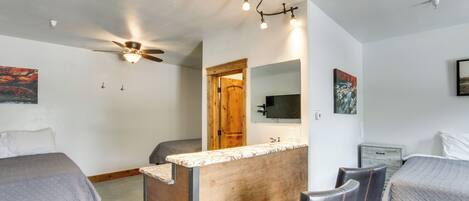 A bedroom with two beds, a wooden desk, a chair, a ceiling fan, and a mounted TV.
