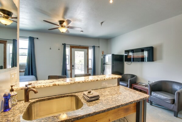 A kitchen with granite countertops, a stainless steel sink, a ceiling fan, and a flat-screen TV mounted on the wall.