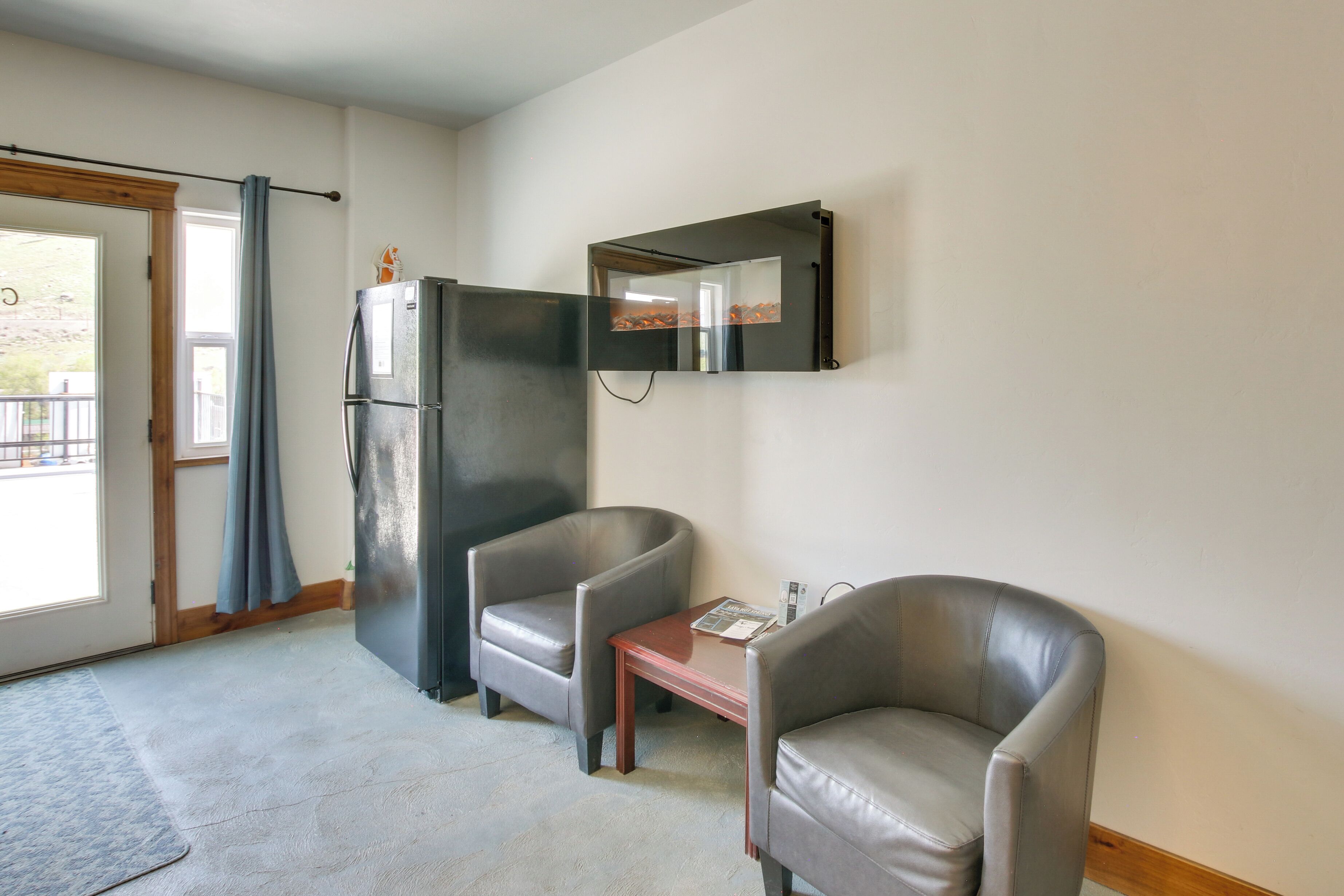 A room with two leather chairs, a small wooden table, a refrigerator, and a mirror on the wall.