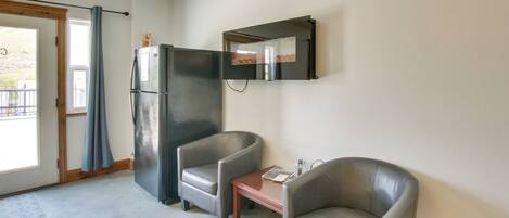 A room with two leather chairs, a small wooden table, a refrigerator, and a mirror on the wall.