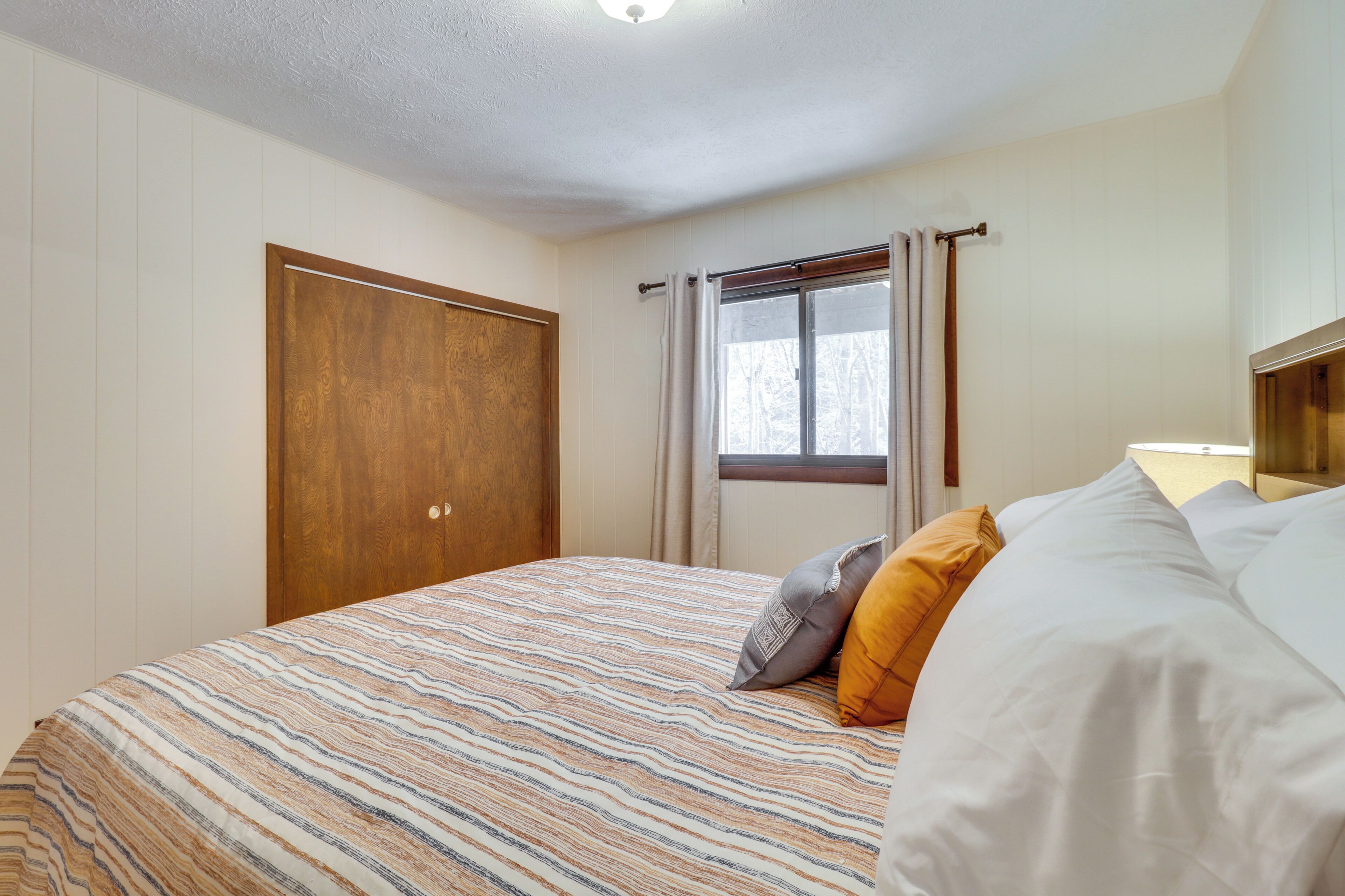 A bedroom with a bed, a wooden wardrobe, a window with curtains, and a view of trees outside.