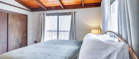 A bedroom with a wooden ceiling, a large bed with white bedding, a window with curtains, and a door.