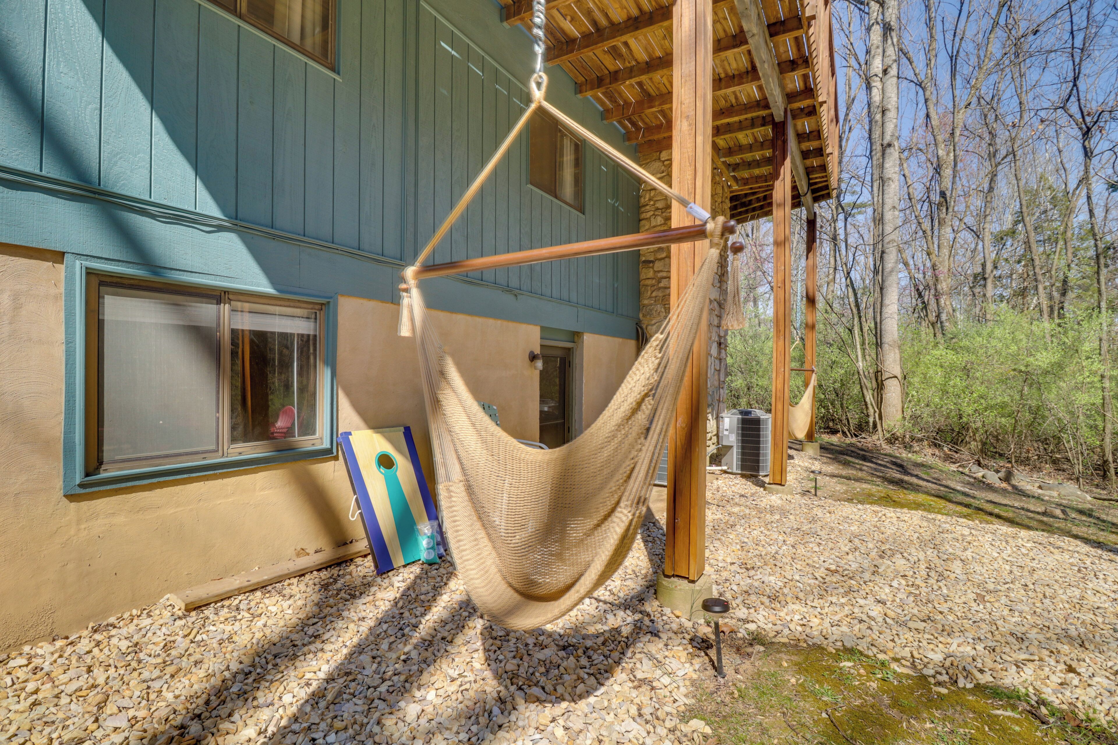 A hammock hanging under a wooden beam, a window with a view of a house, and a gravel path.