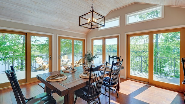 A dining area with a wooden table, chairs, and a hanging light fixture. Large windows provide views of the outdoors. - Lakefront Vacation Rental w/ Views & Hot Tub! (Bracey)