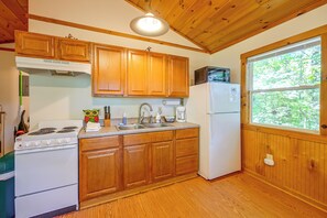 A kitchen with wooden cabinets, a stove, a refrigerator, and a window with a view of greenery. - Cabin w/ Hot Tub - Near Nantahala Outdoor Center! (Bryson City)
