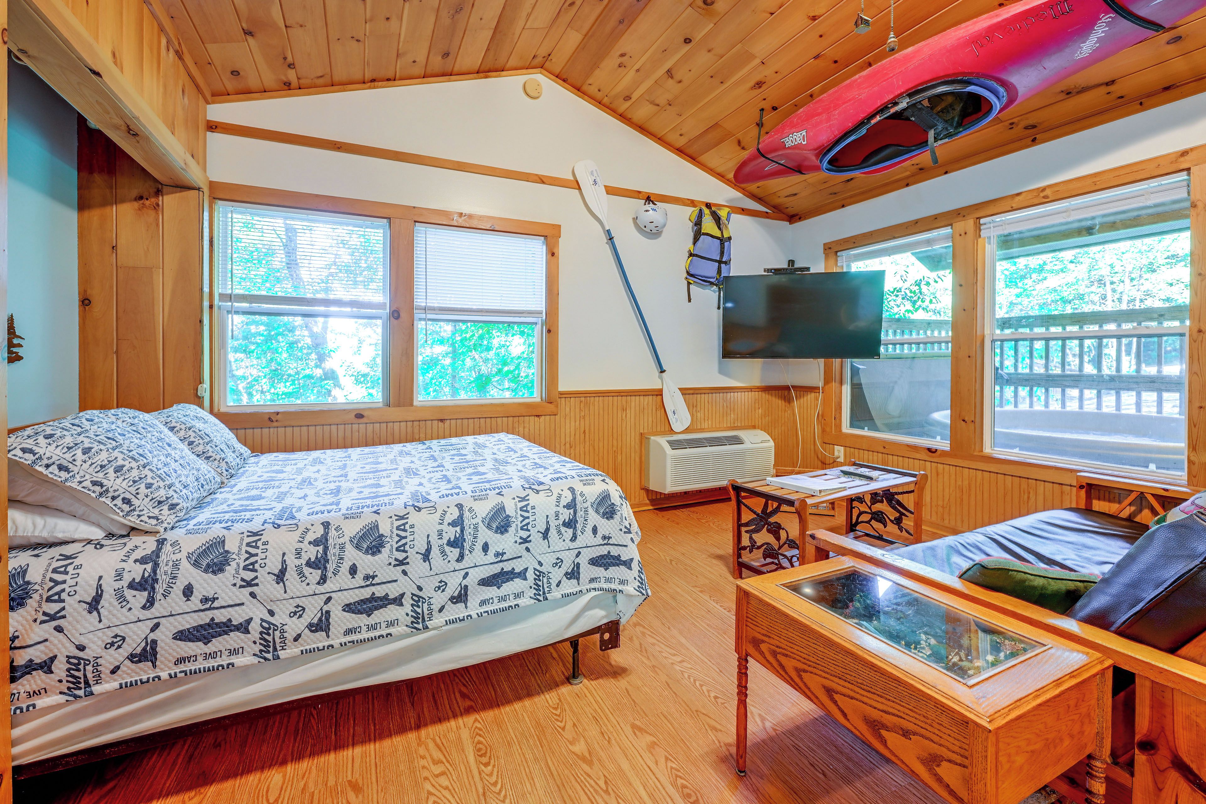 A cabin with a bed, a TV, a table with a glass top, and a kayak.