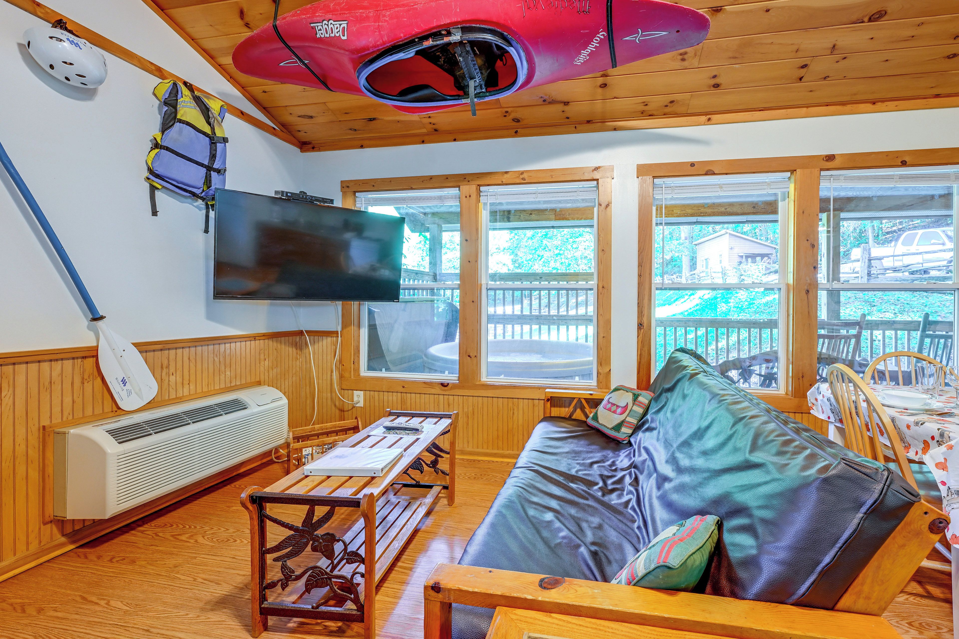 A cabin interior with a wooden ceiling, a mounted television, a table with a laptop, and a couch with pillows.
