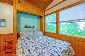A wooden cabin bedroom with a bed, a small stool, and a window with a view of greenery. - Cabin w/ Hot Tub - Near Nantahala Outdoor Center! (Bryson City)