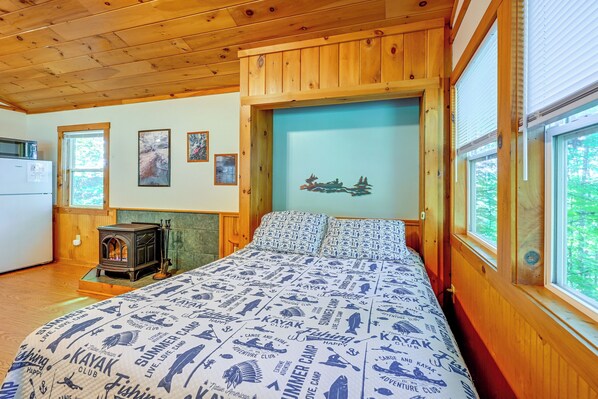 A cabin interior with a bed, a wood stove, a refrigerator, and a window with a view of trees.