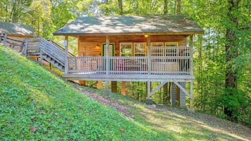 A wooden cabin with a balcony, surrounded by trees and a grassy slope.