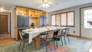 Un coin repas avec une longue table dressée pour six personnes, entourée de chaises. Un ventilateur de plafond est installé au-dessus, et une fenêtre avec des stores se trouve à droite.