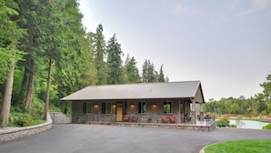 A lodge with a stone facade, a covered porch with seating, and a view of a lake surrounded by trees. - Luxurious Waterfront Retreat w/ Private Pond! (Ridgefield)