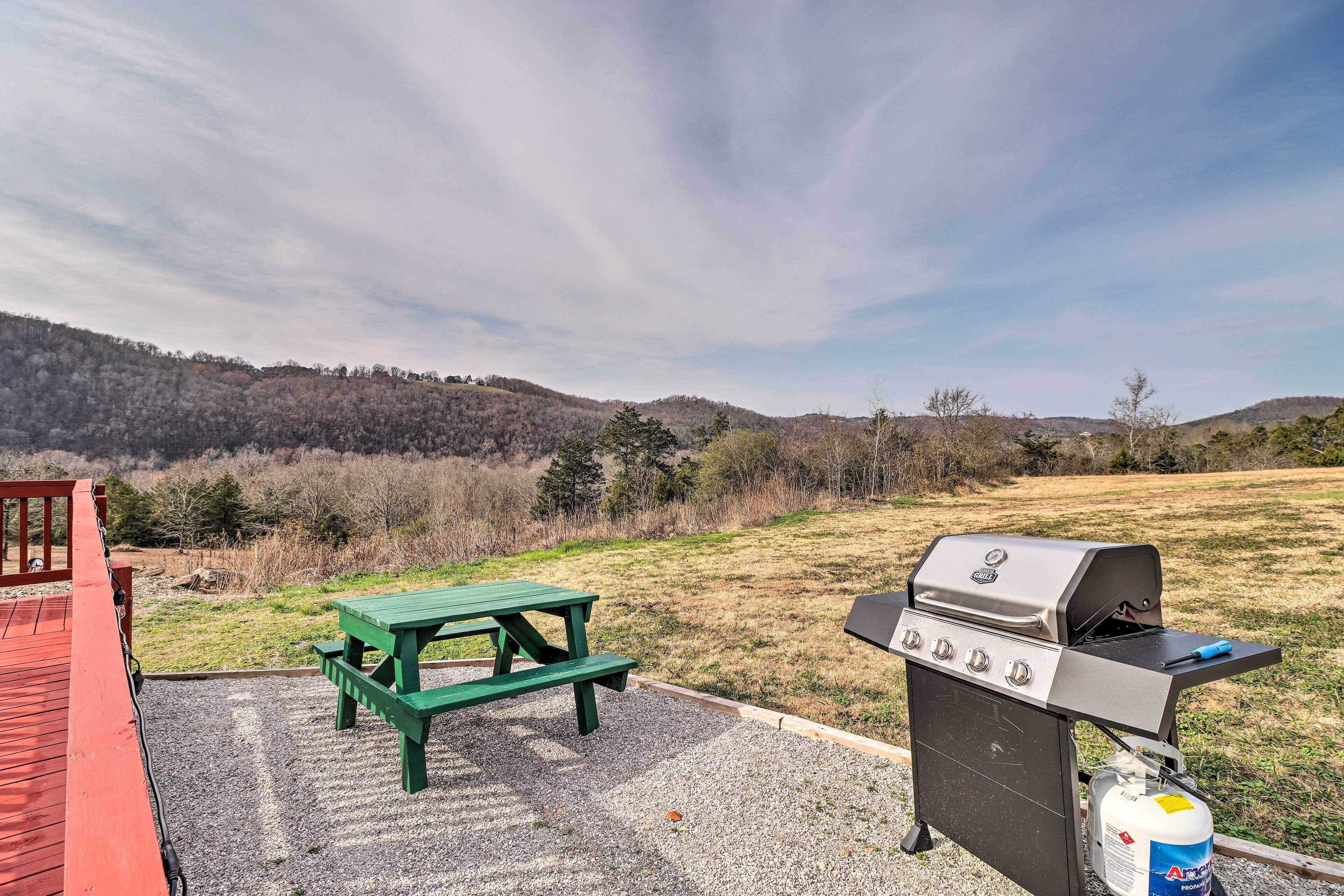 A barbecue grill with a picnic table and a gas canister, set against a backdrop of hills and a clear sky.