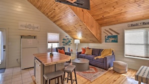 L’intérieur chaleureux d’un chalet avec un plafond en bois, un coin cuisine doté d’un réfrigérateur, une table à manger avec des tabourets, un canapé avec des coussins et une fenêtre avec des stores.