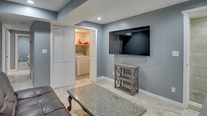 Un salon avec un canapé en cuir, un téléviseur à écran plat fixé au mur et un coin buanderie équipé d’un lave-linge et d’un sèche-linge.
