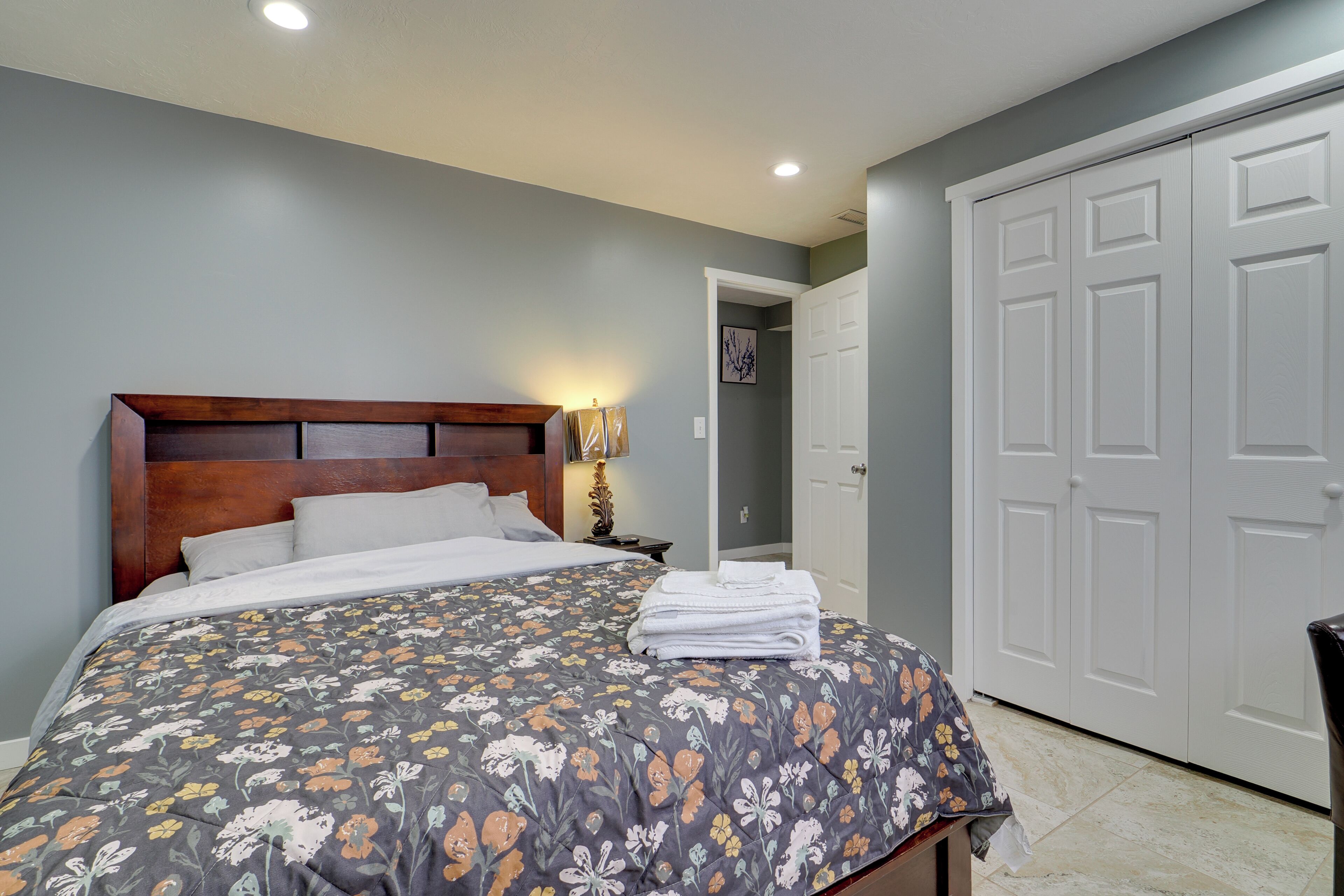 Um quarto com cama, mesa de cabeceira, abajur, porta e corredor.