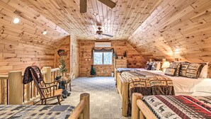 A log cabin bedroom with a high ceiling, wooden walls, and a central window. There are two beds with patterned bedding, a wooden chair, and a small Christmas tree. - Phelps Cabin on 26 Acres w/ Fire Pit! (Phelps)