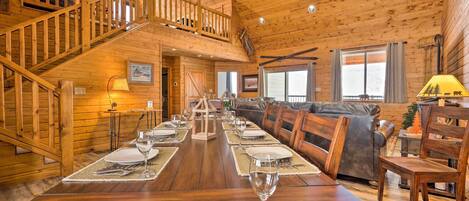 Interior de una cabaña de madera con una mesa de comedor dispuesta para cuatro personas, una escalera y una sala de estar con sofá y sillones.
