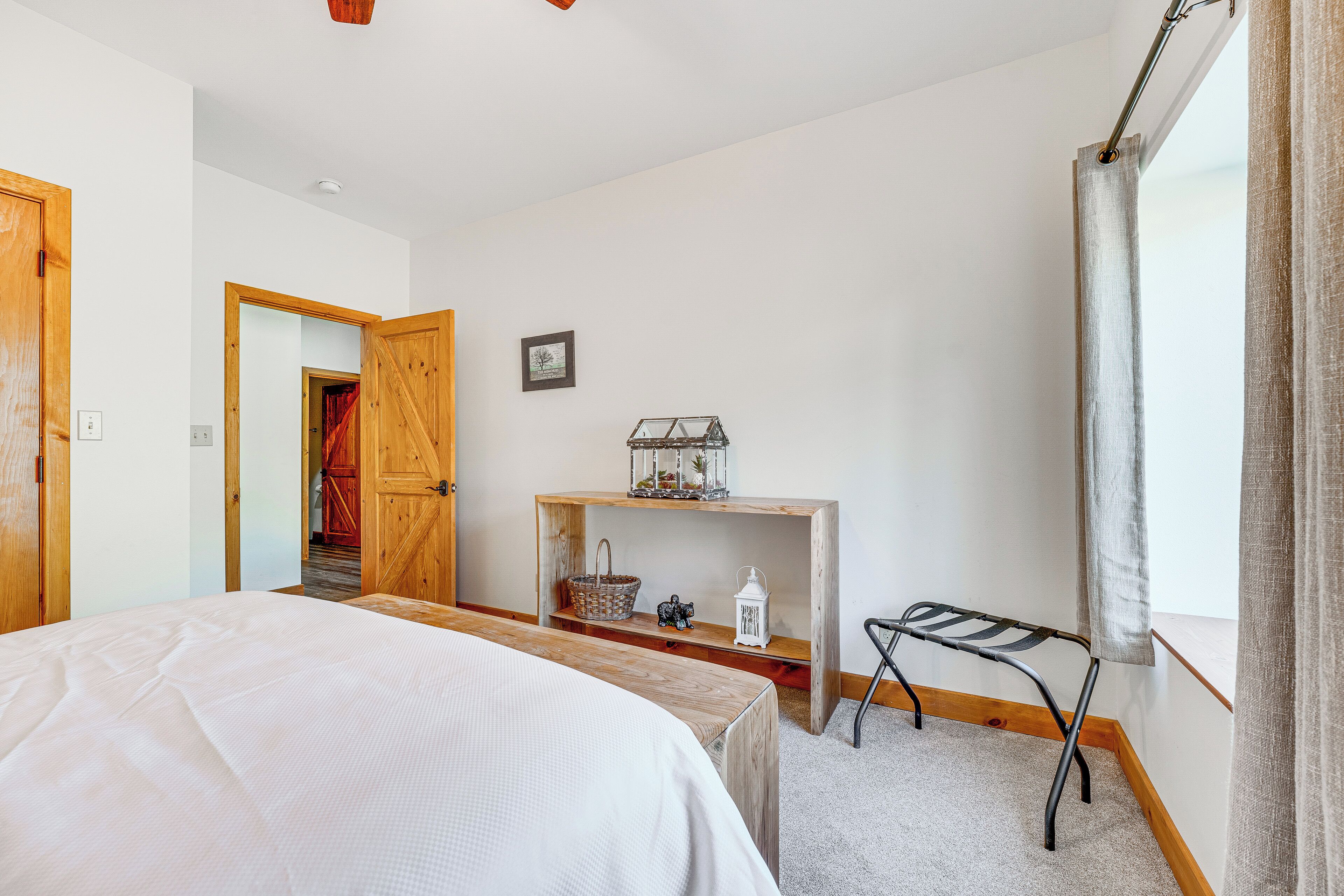 Ein Schlafzimmer mit einem Bett, einem Holztisch, einer kleinen Ablagefläche mit Dekoration und einem Fenster mit Vorhängen sowie einer Tür zu einem anderen Raum.