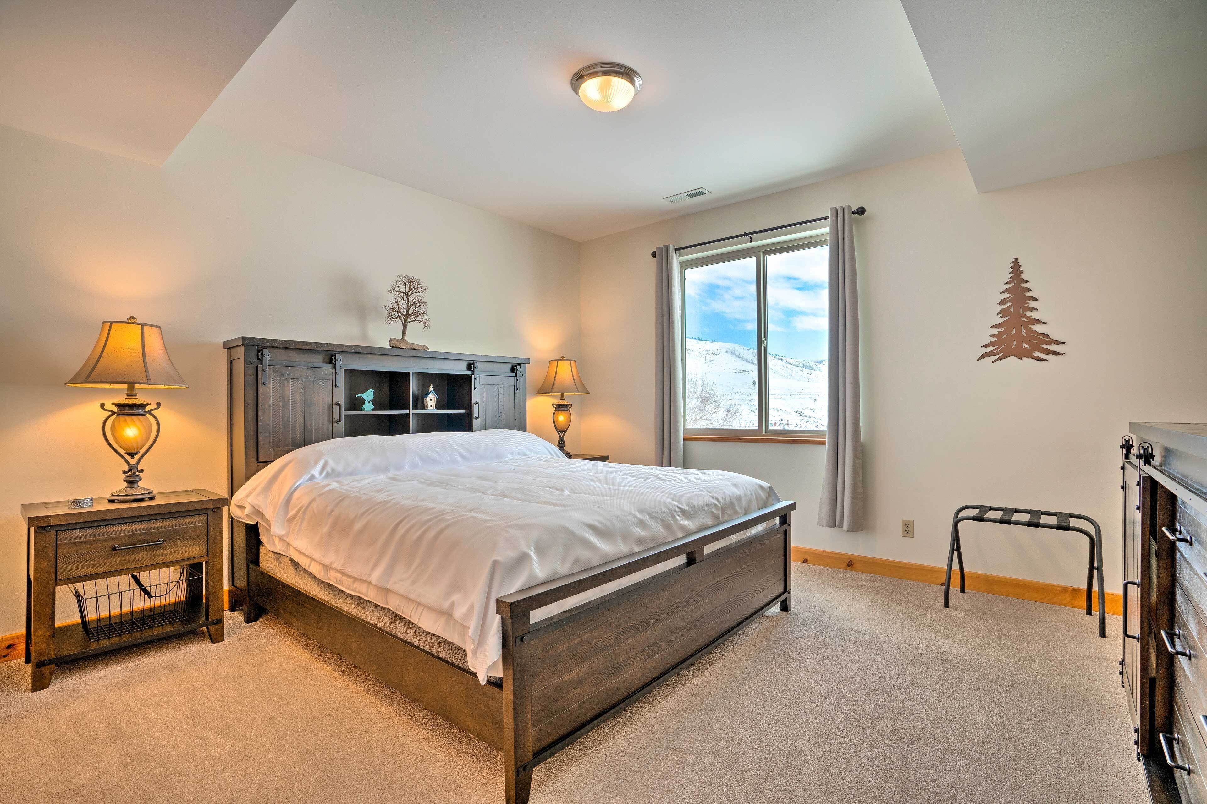 A bedroom with a bed, two bedside tables with lamps, a dresser, a window with a view, and a wall decoration.