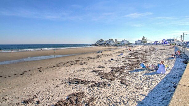 Ein Strand mit blauem Himmel, sandigem Ufer und Gebäuden im Hintergrund.