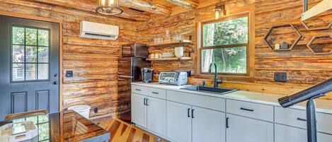 A rustic cabin interior with a wooden interior, a kitchen with white cabinets, a dining area with a glass table, and a large window offering a view of trees.