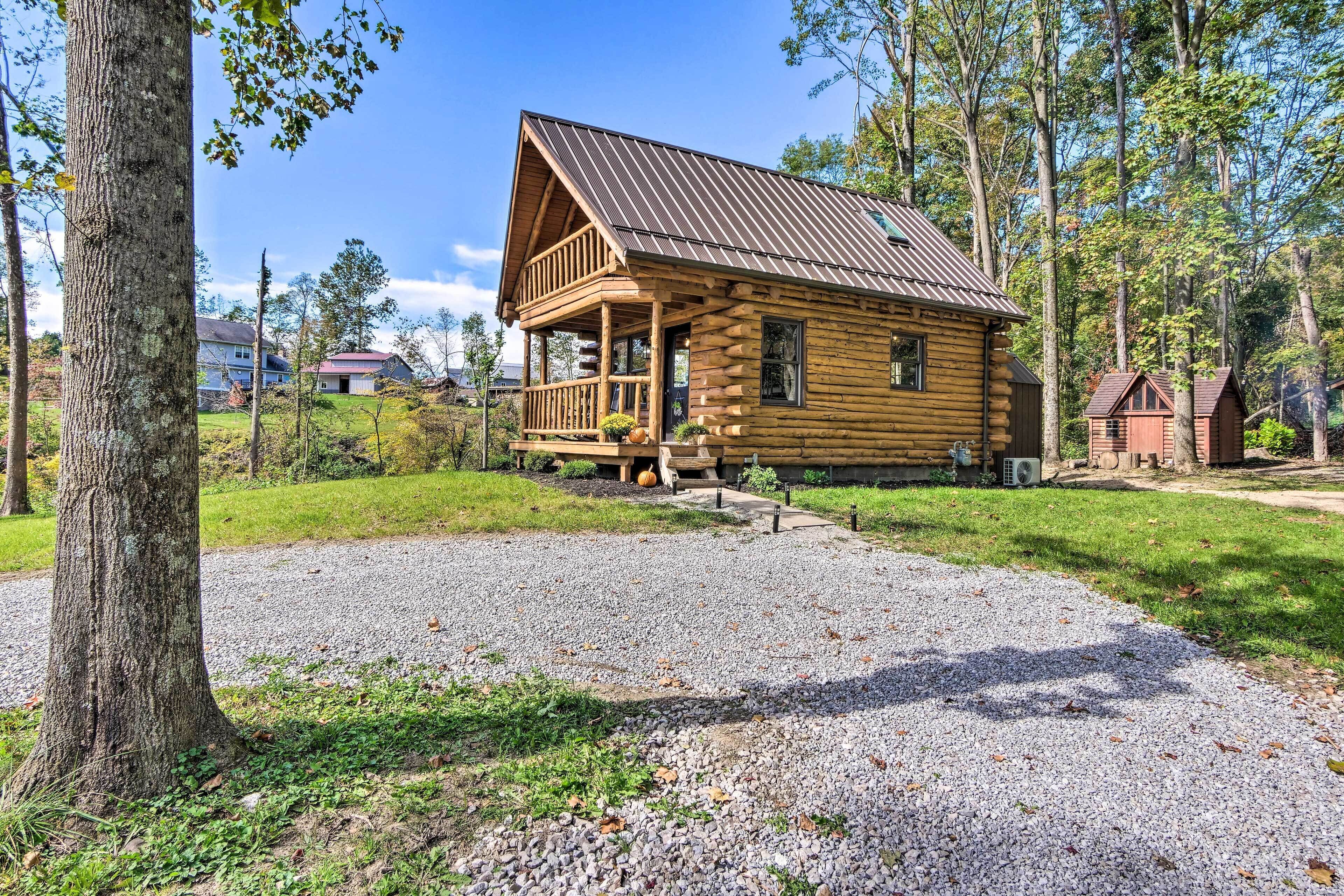 A log cabin with a porch, surrounded by trees and a gravel driveway.