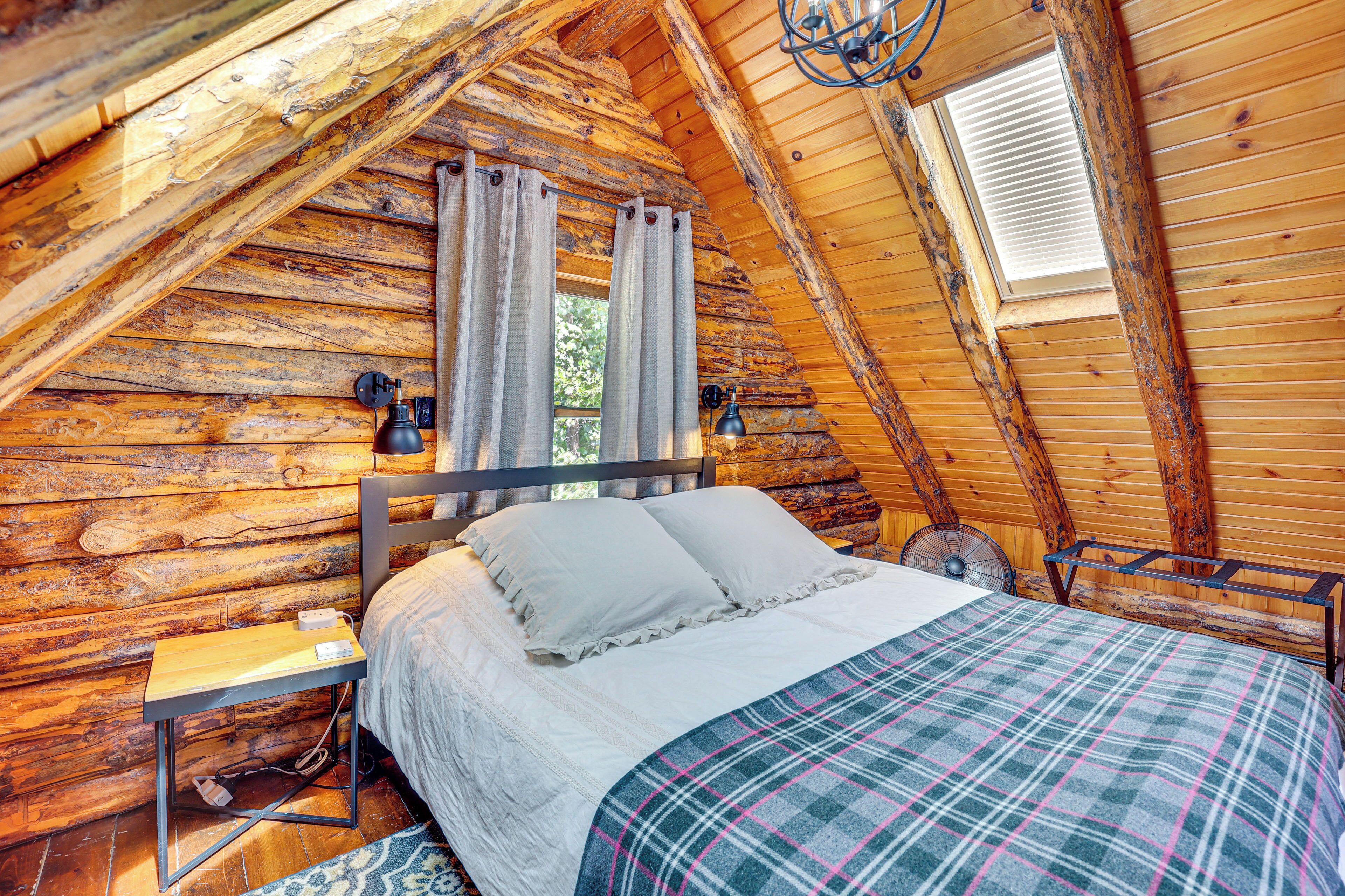 A cozy log cabin bedroom with a wooden ceiling, a bed with white bedding, a plaid blanket, and a nightstand with a lamp.