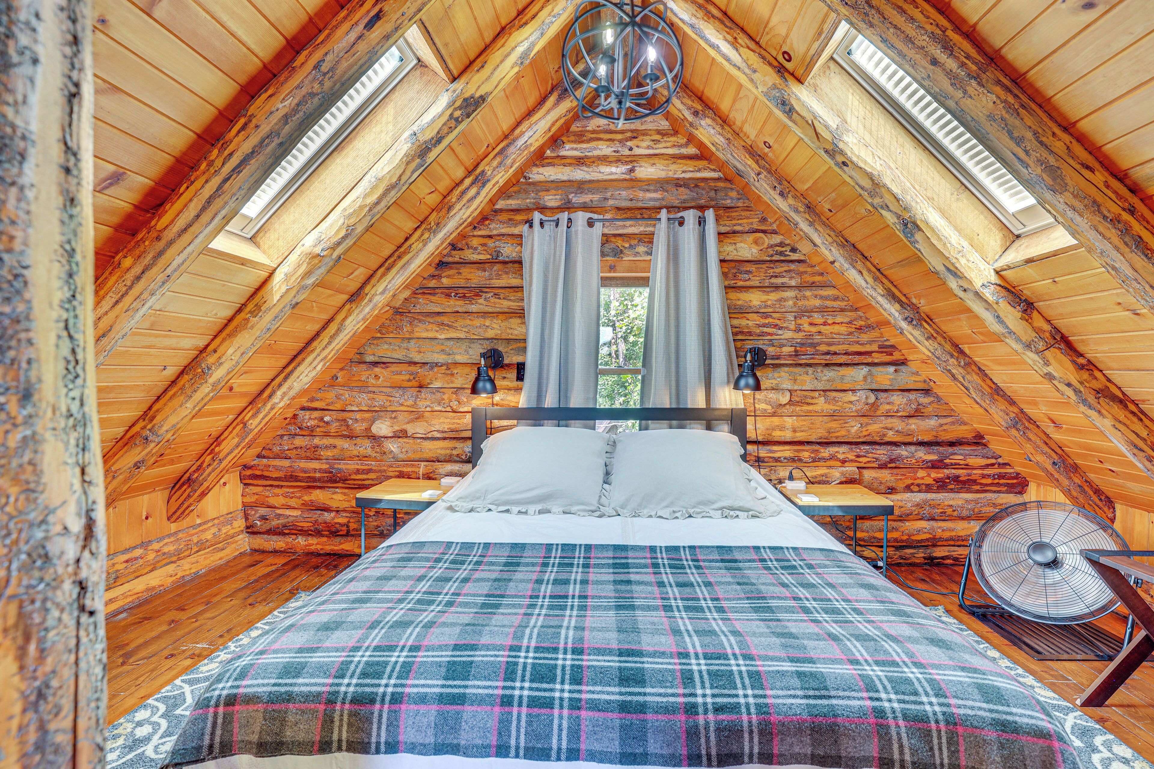 Une chambre mansardée chaleureuse avec un plafond en bois, un lit recouvert d’une couverture à carreaux, deux tables de chevet, une commode et un ventilateur fixé au mur.
