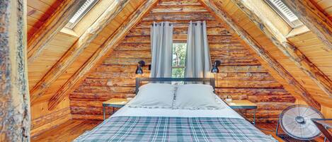 Un dormitorio acogedor en el ático con techo de madera, una cama con manta escocesa, dos mesitas de noche y un ventilador de pared.