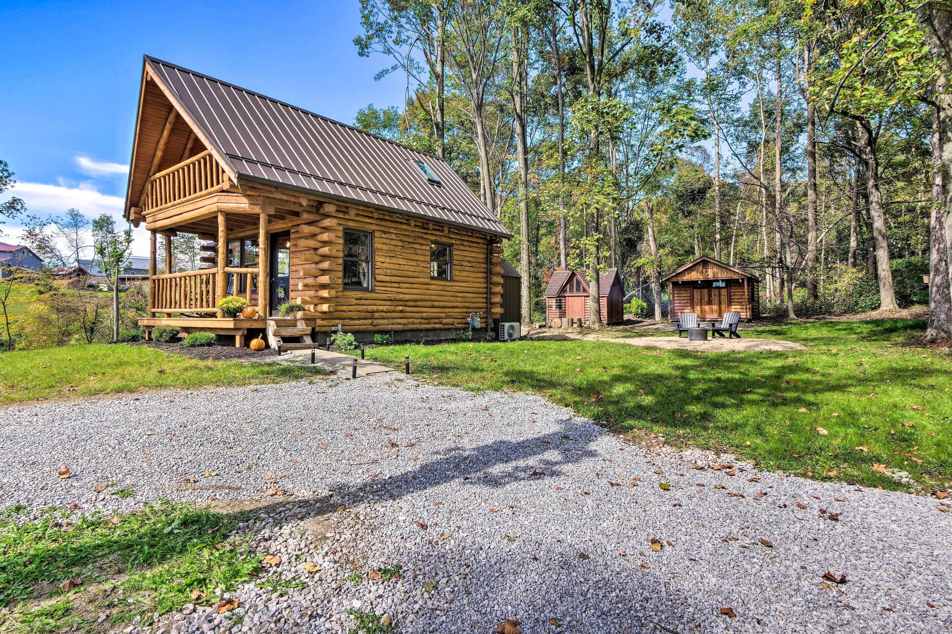 A log cabin with a porch, a small shed, and a gravel driveway surrounded by trees.