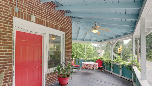 A covered porch with a red door, a potted plant, a table with chairs, and a view of greenery.