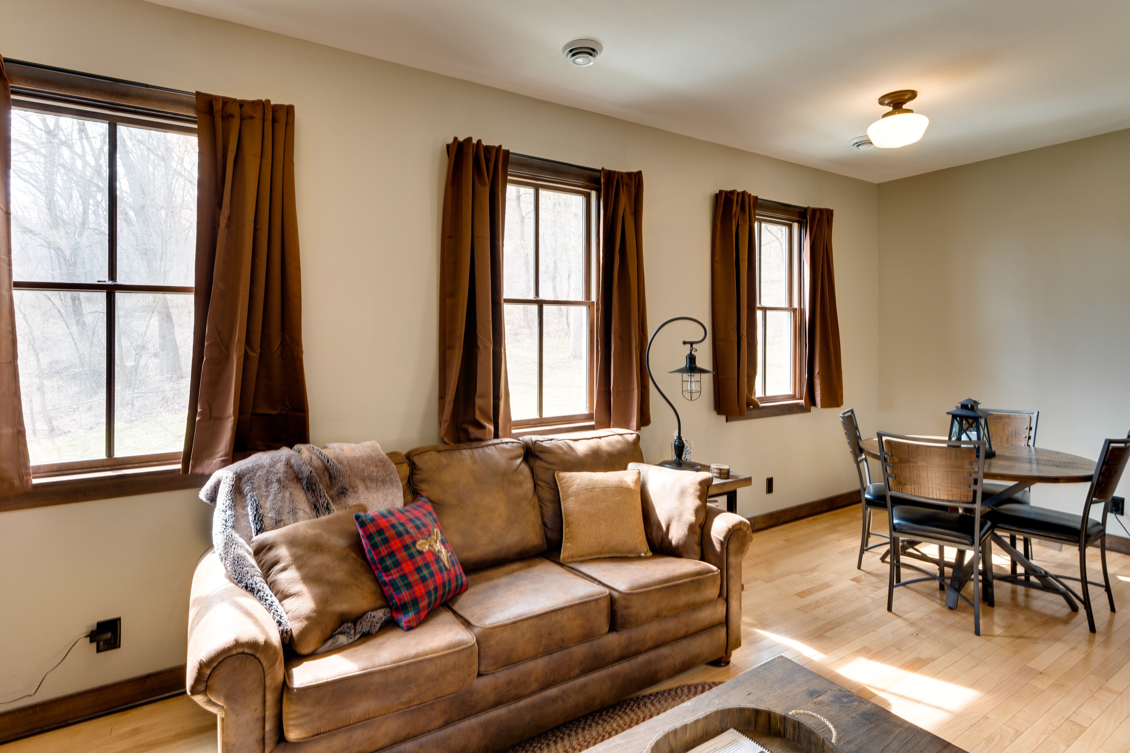 A living room with a brown leather sofa, a dining table with chairs, and large windows with brown curtains.