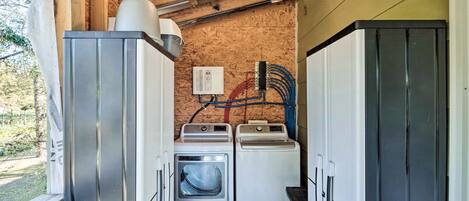 Une petite buanderie équipée de deux machines à laver et d’un sèche-linge, avec des poutres en bois au plafond et des câbles électriques apparents.