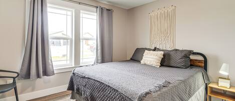 Un dormitorio con una cama grande, suelo de madera, ventana con cortinas, mesita de noche con lámpara y una silla.