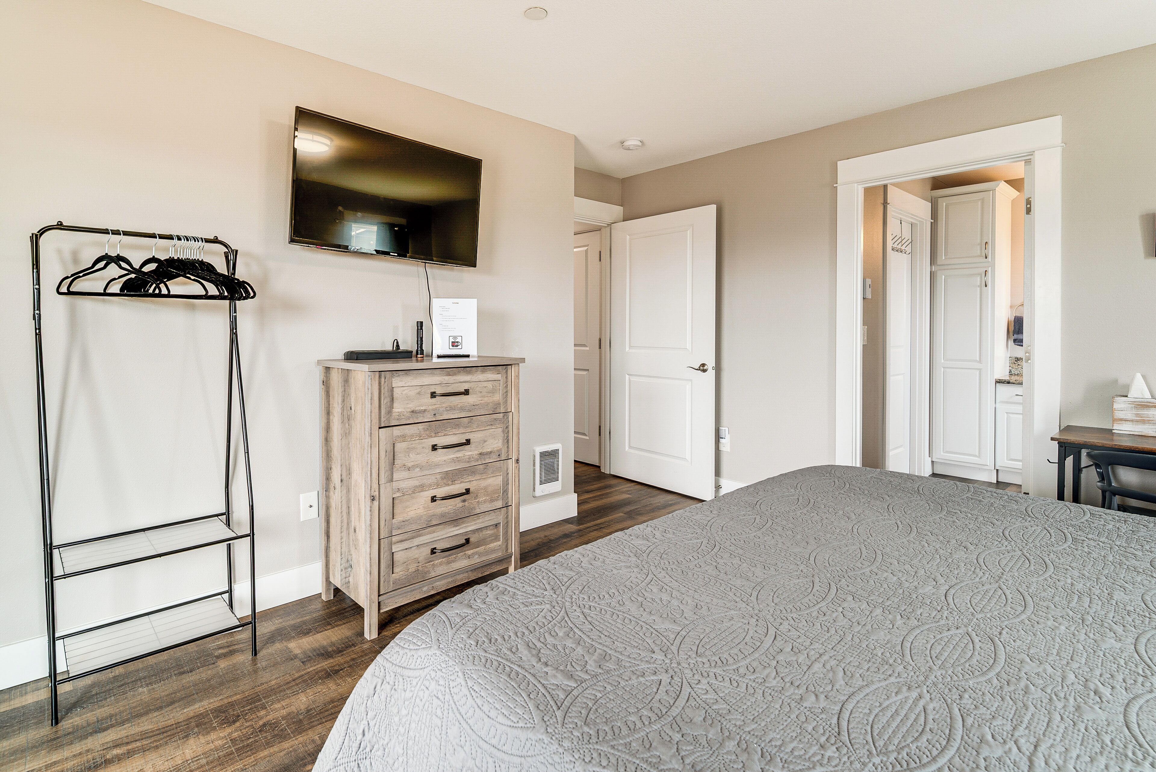 A bedroom with a bed, a wooden dresser, a TV mounted on the wall, and a door leading to another room.