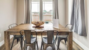 A dining area with a wooden table, four chairs, a decorative centerpiece, and a view through a window with curtains. - Modern Ocean Shores Home, 2 Mi to Beach! (Ocean Shores)