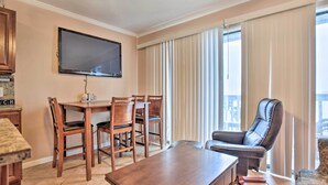 A living room with a wooden dining table, four chairs, a flat-screen TV mounted on the wall, a leather recliner, and large windows with vertical blinds. - Oceanfront Corpus Christi Condo w/ Pool! (Corpus Christi)
