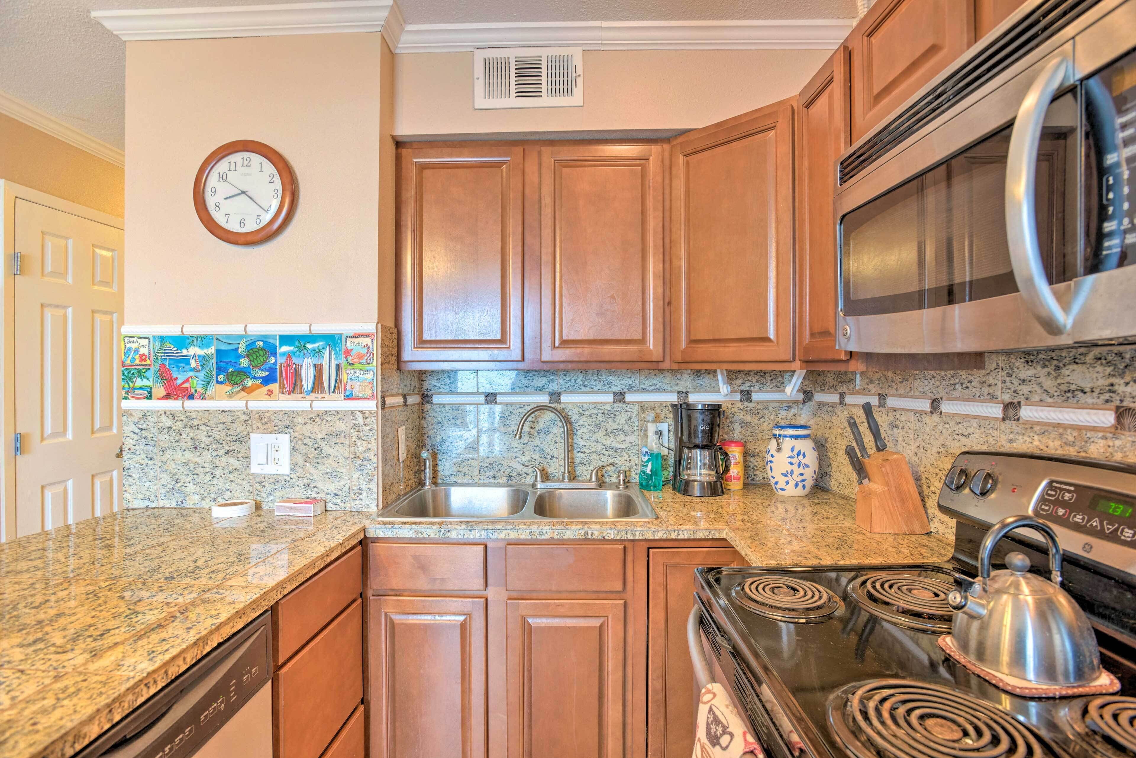 A kitchen with wooden cabinets, a stainless steel microwave, a stove with a kettle, a sink, and a clock on the wall.