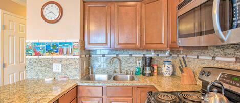 A kitchen with wooden cabinets, a stainless steel microwave, a stove with a kettle, a sink, and a clock on the wall.